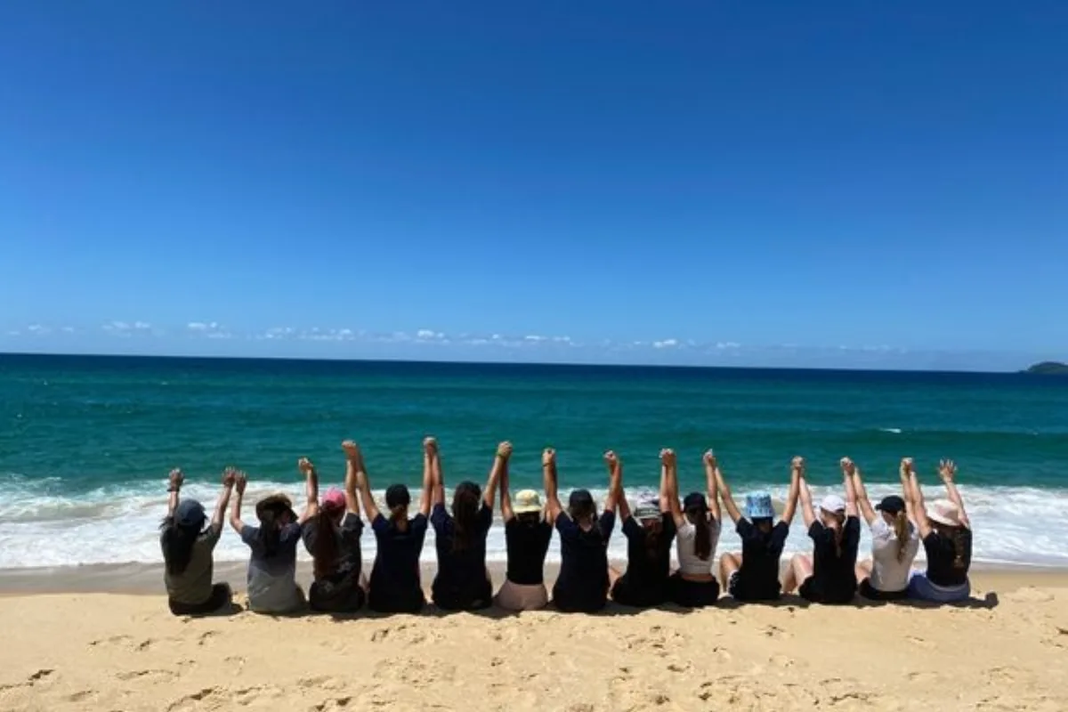 Students sitting together on a beach with arms raised after completing a journey